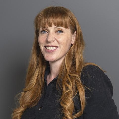The image shows Angela Rayner with long, wavy, reddish-brown hair, wearing a dark-colored button-up shirt. She has a warm smile and is looking directly at the camera. The background is a plain, neutral gray, which contrasts with her hair and clothing. The lighting is soft and even, highlighting her features without creating harsh shadows. Her hair is styled with loose curls, and she has bangs that frame her face. The overall composition is simple and focused on the subject, with no additional objects or distractions in the frame.

@altbot, edited by @jemmesedi