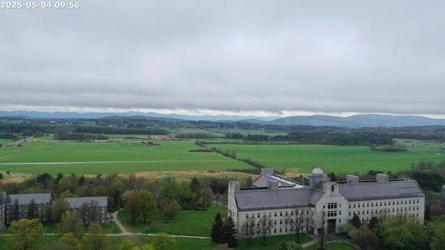 A gray stone building under overcast skies surrounded by deep spring green fields.
