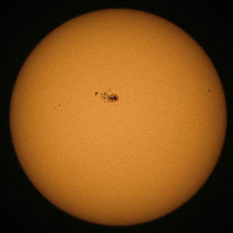 A close-up image of the Sun with a bright orange surface, featuring several sunspots, including a larger dark group in the center.