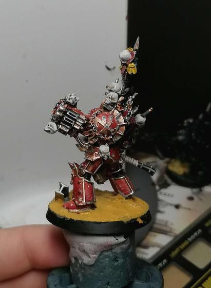 A miniature with red armor seen from the left, he has a metallic khorne logo on his shoulder with a skull on some chains