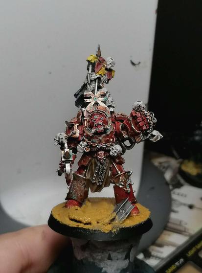 Sam miniature seen from the front, he has red and gold armor with a spike with an empalled comissar on it on his back. His skull has a grinning/screaming mouth with teeth.
He has an axe in his right hand and a bunch of skull in his left.