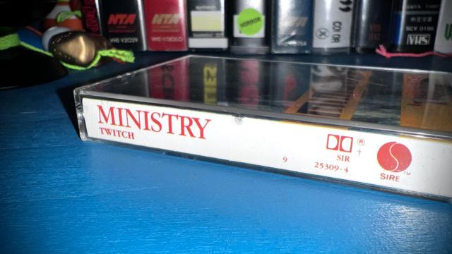 A cassette tape of Ministy’s “Twitch” sitting atop a blue end table. The spines of several vhs tapes are visible in the background.