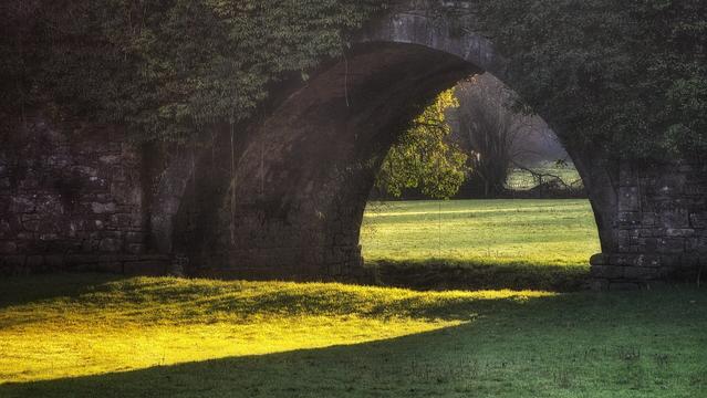 Sunlight illuminates green grass under the stone arches of an old bridge, framed by trees.
