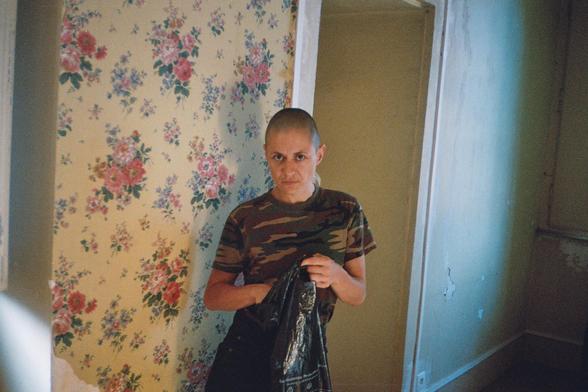 Gorge en t-shirt camouflage tenant un veste dans un sky dans les mains, elle pose dans un couloir à la tapisserie vétuste.