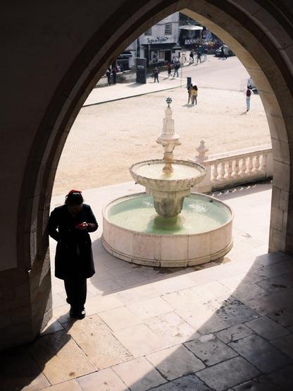 A woman uses her phone under a tall arch. Besides her is a fountain and bright sunlight, while on the near side of arch is darkness.