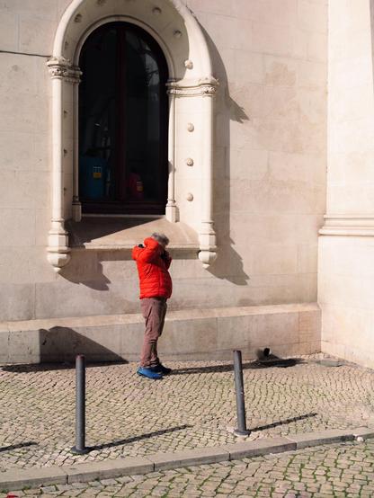 A man with a red jacket stands near a wall and a decorated window with an arch, seemingly talking to the phone. He has one hand on the back of his head and the other hidden, probably holding his phone.