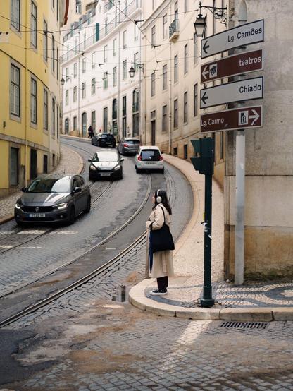 A young woman waits at an intersection as cars come and go from a pebble street that goes up a slope between tall buildings with regular windows.