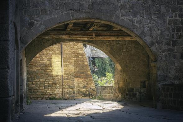 The historic San Pelligrino quarter in Viterbo Italy contains an “interesting variety of 11th to 13th century architecture”. The result is a pleasing array of lines, angles, and arches. Here is seen bright sun creating interesting interplay of light and shadow on a cobblestone walkway underneath a stone arch with a peaceful green garden area beyond.