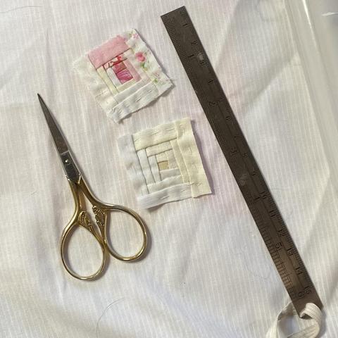 Two tiny log cabin pattern patches the upper one on a pink-white colourway and the lower one is a cream-white colour contrast so very similar but it still shows in the patch. Thee is a mini craft ruler to the right and a pair of small embroidery scissors to the left. The patches are one and a sixth of an inch square.
