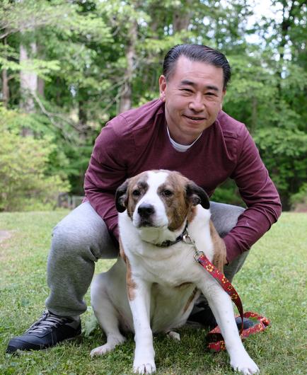 A man in maroon shirt crouching in the yard holding his white and brown dog in the front, not cooperating...
