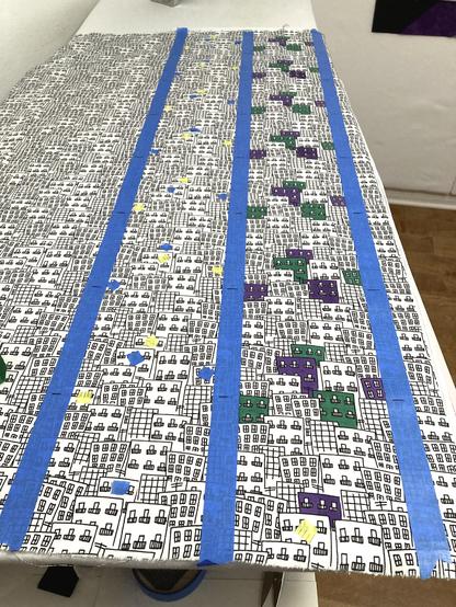 A swath of fabric laid out neatly over a long table. The fabric design is overlapping hand-drawn buildings resembling a crowded city. Three pieces of blue painters tape run from top to bottom, making two columns. The rightmost column has randomish buildings painted green and purple. The leftmost column has small bits of blue and yellow tape where green and purple paint will go tomorrow.