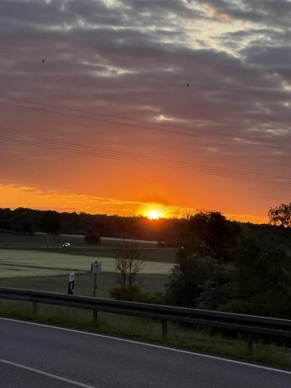 Ein stimmungsvoller Sonnenaufgang über einer ländlichen Landschaft. Die Sonne erhebt sich am Horizont hinter einer Baumreihe und taucht den Himmel in leuchtendes Orange und Rot, während darüber dunkle Wolken dramatisch den Himmel durchziehen. Im Vordergrund führt eine Landstraße mit Leitplanke durch sanfte Felder, auf denen sich der neue Tag langsam entfaltet. Ein Auto ist in der Ferne unterwegs - Symbol für den beginnenden Alltag im Licht eines friedlichen Morgens.
