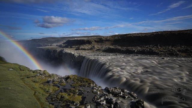 The image shows a powerful waterfall cascading into a gorge, with a rainbow visible to the left, arcing down toward the water. The scenery includes rocky cliffs and a grassy area in the foreground. The sky is blue with some clouds.