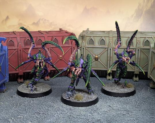 3 Tyranid Alien creatures with huge claws and tentacled faces. They have purple flesh and striped green carapace. They stand amongst shipping containers on an alien world.