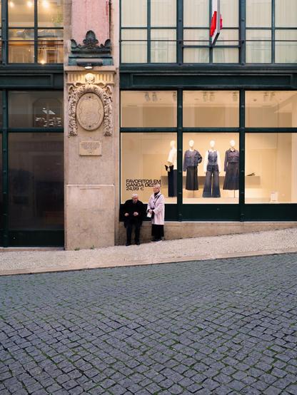 An elder couple rests in front of a clothes shop window. The man is sitting on the stone base of the window, while the woman is standing.