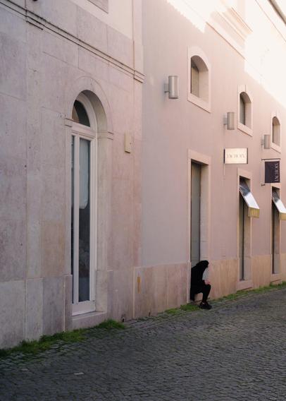 A yound woman wearing black clothes is sitting on a door sill while using her phone and smoking. Bright sunlight comes from above, but the alley is in the shade.