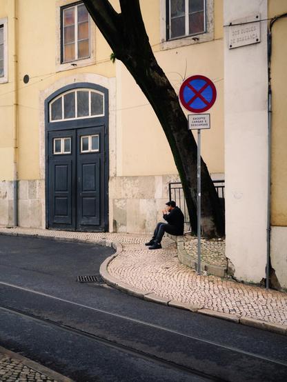 A man with a had lights a cigarette while sitting on a stone step beneath a tall tree. Beyond him there's a building with a large black gate.