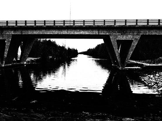 A road bridge crosses the calm water of a canal. In this high contrast black and white image not much detail is seen in the distant trees and landscape.