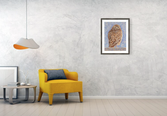 A watercolor painting of an owl is hung on a wall near a yellow chair, lamp and table.