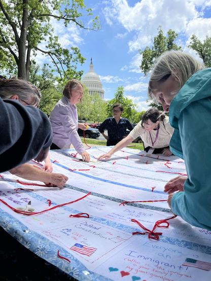 A group of women hand tying a quilt on a frame. The women are outside under a tree and the US Capitol building can be seen behind them.