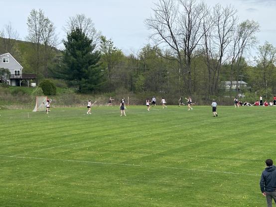 A lacrosse game is being played on a grassy field. Players in black and white uniforms are positioned near the goal, with a referee overseeing the game. In the background, there are trees and a house visible. Spectators are seated on the sidelines.