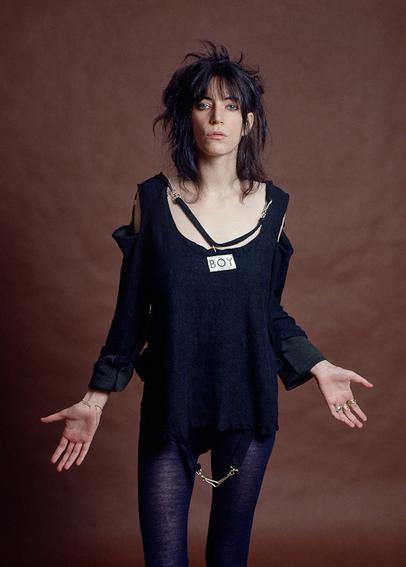 A person wearing dark blue and denim, with dark hair, with their arms outstretched, and wearing a tag that reads, "BOY".
