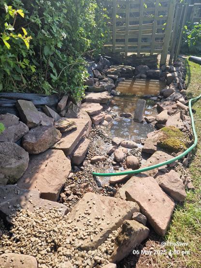 The picture shows a long narrow pond being filled.
Large sandstone rocks surround the pond, with a shallow gravel area closest to the camera