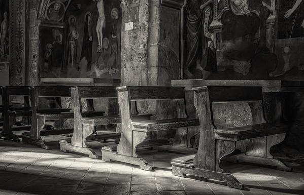 Sunlight spills in through a window and onto some of the wooden pews of the church of Santa Maria Nuova, a Romanesque church in Viterbo Italy. Murals depicting religious figures adorn the stone walls, adding a sense of reverence and antiquity to the scene.  Processed in BW.