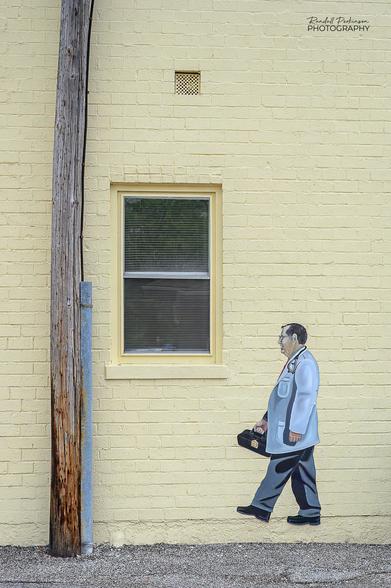 A brick building painted yellow with a window, a vent, and a graphic of a doctor wearing a white lab coat and carrying a medical bag.  A telephone pole is located directly adjacent to the building.