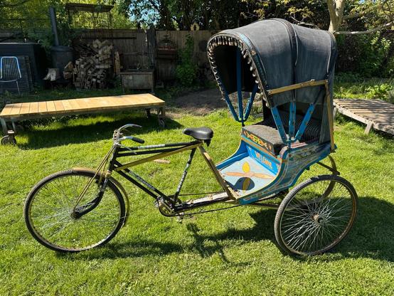 In einem Garten steht eine Fahrrad-Rikscha, mit silbernem Rahmen, blauem Boden und grauer Markise über dem Mitfahrer-Abteil am Heck.