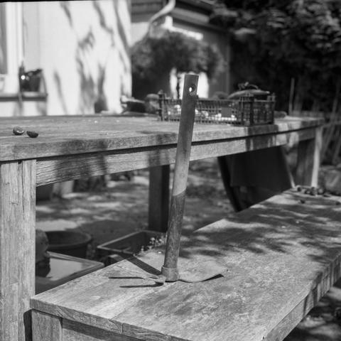 Black and white photo of a rustic wooden table and bench outdoors, with a garden hoe placed upright on the surface of the bench. In the background, various gardening tools and a crate are scattered around, with a house in the background and foliage casting shadows across the scene