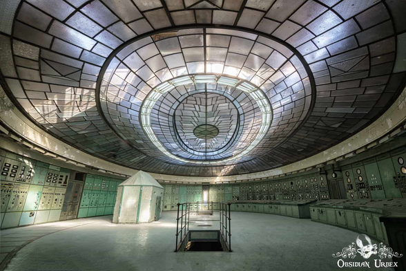 art deco control room, oval with immense skylight. Stairs lead down, there is a curved control panel on the right covered with dials and switches (the walls on both sides are similarly covered). There is a concrete bunked to the center left