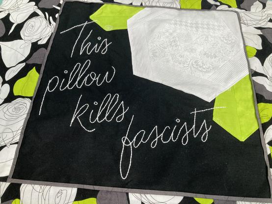 Hand embroidered text, white on a black background, This pillow kills fascists. There’s a pieced white rose design in the upper right and some white scribble rose fabric showing, but the full pillow top isn’t visible