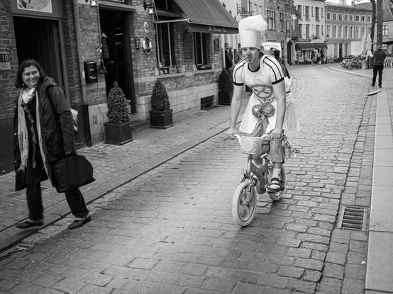 The image is a black and white photograph depicting a street scene in Bruges, Belgium. In the foreground, a person is riding a small bicycle, wearing a chef's hat. The bicycle has a basket at the front. The rider is wearing sandals and appears to be in motion, pedaling down the cobblestone street. To the left, a woman is walking, carrying a bag and wearing a scarf and a dark coat. The background features brick buildings with storefronts. The street is lined with potted plants, and there are other pedestrians in the distance. The overall atmosphere is lively, with a sense of movement and everyday life.