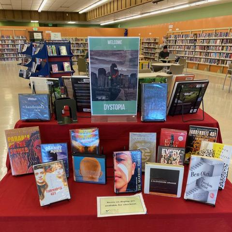 A display of books and videos that all have dystopian themes. The poster in the middle says "Welcome to Dystopia", with "You are Here" in small letters.