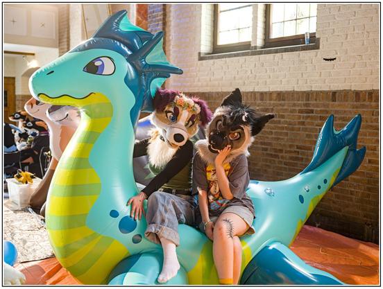 Two fursuiters (one light colored with floppy ears like a collie, the other looking more like a hyena) riding a big blue inflatable sea dragon