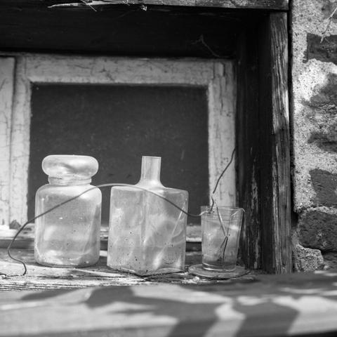 Black and white Photo of an old cookie jar, a vase and a water glass on the shelf of an old window with a wooden frame in a brick wall.