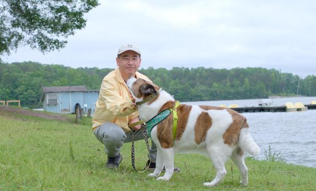 A man with a white cap and a yellow jacket crouching on the grass by a lake, holding his white and brown dog. A light blue old boat house on the left, and a deck with paddle boats on the right.