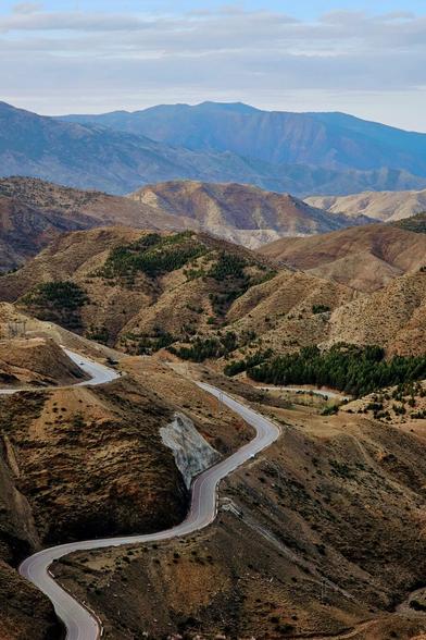 A high-angle shot shows a long, winding paved road cutting through a vast, hilly, and mountainous landscape under a partly cloudy sky.