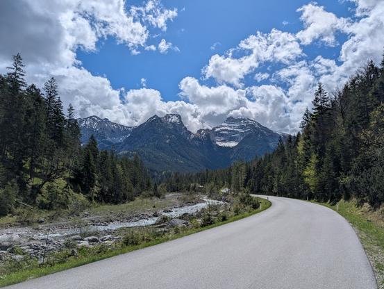 Straße neben dem Rissbach. im Hintergrund hohe Berge. Einige Wolken am blauen Himmel.
