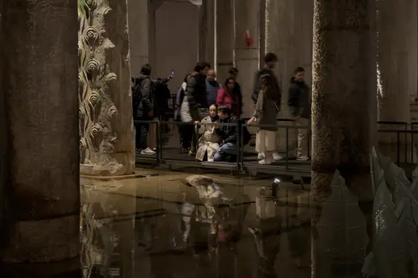Photograph of man and a woman inside an ancient underground water reservoir. They are kneeling, focused on reading the museum label for one of the ancient columns surrounding them. The column surrounding them is engraved with swirls, whereas the other columns in the photograph have no such decorations. They are surrounded by other tourists, but the others are blurred by movement. The visitors are all on a metal platform elevated slightly above the shallow water in the room. The pillars and the tourists, including the man and the woman reading, are reflected in the water.