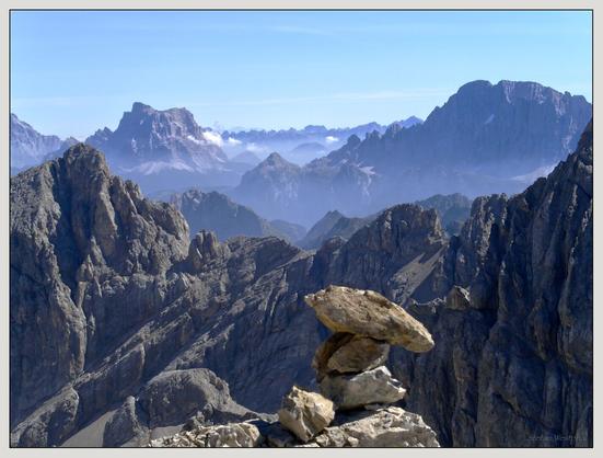 Ein felsiger Gipfel im Vordergrund mit einem ausbalancierten Steinstapel, der mehrere Reihen ausgedehnter Gebirgsketten mit blauen und grauen Schattierungen unter einem klaren Himmel überblickt.

A rocky peak in the foreground with a balanced stack of stones overlooking several rows of expansive mountain ranges with shades of blue and gray under a clear sky.