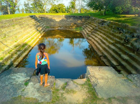 Photo shows a water basin with stairs sides of appx 3 meter with eleven steps until the water level. In front there is a young girl with her back to the photographer to show the scale of the construction. She is appx 1,2 meter tall, have her long hair in a ponytail and has a sleeveless very colourful dress. Around her waist she has draped a deep blue hoodie. She is wearing pink sandals. It is surrounded by green turf and then bordered by trees. Through the trees some distant hills are shown, telling of this basin belonging to a mountain temple complex. The sky is partly clouded and a bit misty.