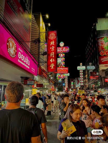 A snapshot from a crowded walkway in Bangkok, people are walking away from and towards the camera, brightly colored signs and building create a perspective shot angling towards the center of the frame, the dark night sky slices downward from the top right of the frame, one man appears to be rubbing his hands together having accomplished a meal, someone wears an Elmo shirt, a women in the front center holds a yellow box of food now eaten