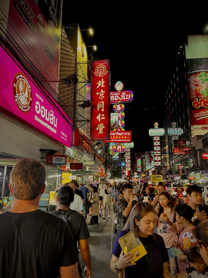 A snapshot from a crowded walkway in Bangkok, people are walking away from and towards the camera, brightly colored signs and building create a perspective shot angling towards the center of the frame, the dark night sky slices downward from the top right of the frame, one man appears to be rubbing his hands together having accomplished a meal, someone wears an Elmo shirt, a women in the front center holds a yellow box of food now eaten