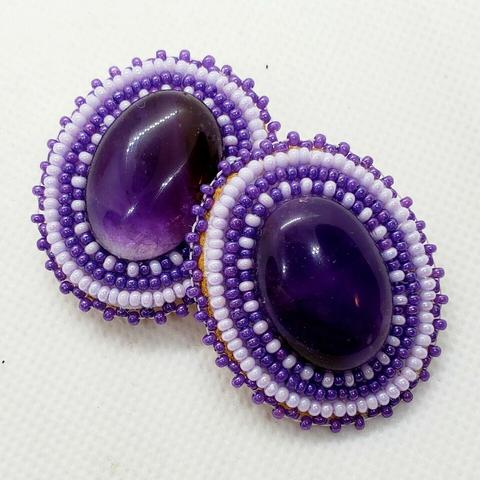 a pair of beaded earrings made by kiamichi. they have translucent deep purple gemstones as centerpieces surrounded by shiny purple and white bead designs