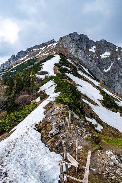 Ein Bergsattel mit einem gekennzeichneten Weg. Es fällt rechts und links ab, es liegt auch auf beiden Seiten stellenweise noch Schnee. Der Weg führt nach oben und endet in schroffen Felswänden.