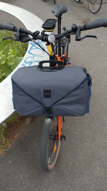 Blick von vorne auf ein Brompton Electric G-Line mit montierter, großer, grauer Fronttasche mit mittig spitz zulaufendem Deckel.