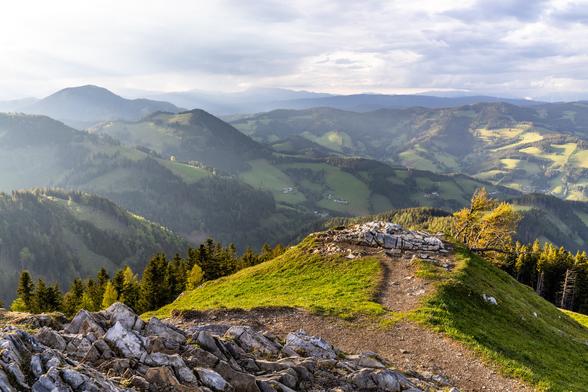 Blick von einem Gipfel in die weite Landschaft, die von der bereits tiefstehenden Sonne bestrahlt wird. Der Himmel bietet hochstehende Wolken. Es ist eine hügelige und bergige Landschaft. Im Vordergrund Fels und ein kleiner Vorsprung.