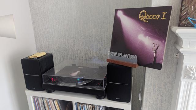 We have a black turntable flanked by two black speakers on some white furniture with grey wallpaper behind. The playing record is black vinyl. On the right speaker are the words 'Now Playing' lit up in front of the record sleeve.

The sleeve shows a white spotlight shining from the top left to the bottom right where a stylised Mercury stands with a microphone stand above his head. The rest of the sleeve contains various hues of purple.